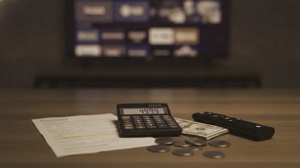A calculator, dollar bills, coins, a streaming stick and a remote on a table in front of a TV