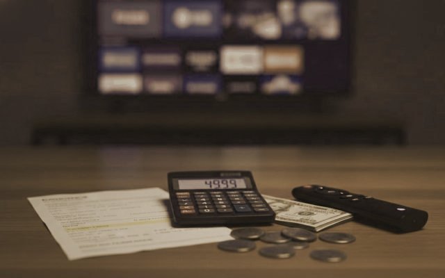 A calculator, dollar bills, coins, a streaming stick and a remote on a table in front of a TV